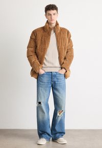 Braune Cord-Pufferjacke, beiger Pullover darunter, verwaschene hellblaue Jeans, hellfarbige Schuhe, lässige Passform, strukturierte Oberfläche.