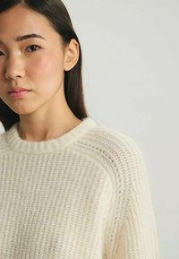 Pull en tricot de couleur crème, avec une texture côtelée et un col rond. Le tissu semble doux et léger, avec des motifs subtils.