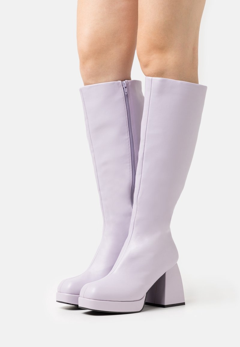 Bottes lavande jusqu'à la jambe avec talons blocs et fermetures éclair latérales portées par une personne debout sur un fond blanc.