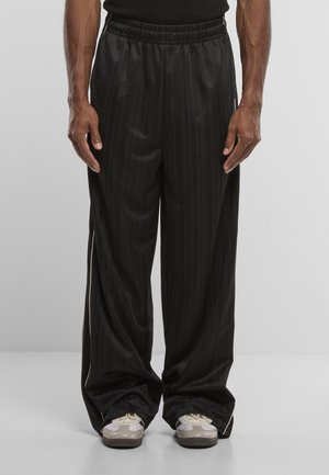 Homme portant un pantalon ample noir rayé et des baskets blanches sur un fond gris uni, mains détendues le long du corps.