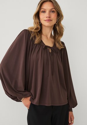 Kvinde med bølget blondt hår iført en løs brun bluse med bindebånd ved halsudskæringen og voluminøse lange ærmer, kombineret med sorte bukser.