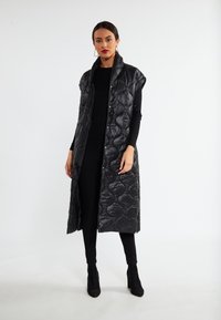 Gilet lungo nero trapuntato con colletto alto, motivo testurizzato, design senza maniche e chiusure a pressione. Indossato sopra un top nero aderente e pantaloni.