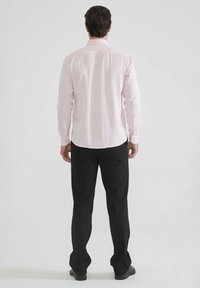 Chemise rose pâle à manches longues avec un ourlet incurvé et des poignets à boutons, associée à un pantalon noir et des chaussures noires. Vue arrière montrant des lignes épurées et une coupe soignée.