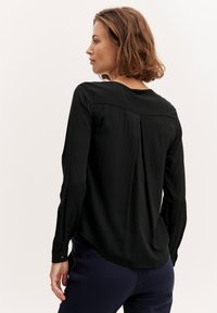 Maglietta nera a maniche lunghe con scollatura tonda, texture morbida, dettaglio a pieghe sul retro e polsini con bottone. Indossata sopra pantaloni blu navy.