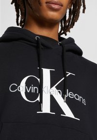 Crna dukserica s velikim bijelim "CK" logotipom i tekstom "Calvin Klein Jeans", s srebrnim detaljima na vezicama i mekanom, teksturiranom tkaninom.