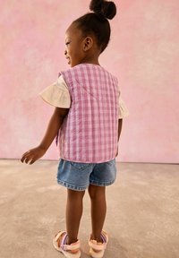 Vadderad väst i rosa rutigt mönster, lager på lager över en vit topp, tillsammans med denimshorts och ljusa sandaler. Bakgrunden är rosa.