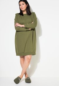 Vestido oversized de color verde oliva con mangas largas y cuello redondo. Tejido suave, de longitud media, combinado con zapatillas verdes con acentos de cadena.