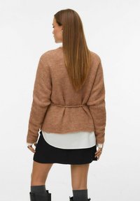 Terakota pleteni kardigan opuštenog kroja, dugih rukava, s vezanjem u struku, nosi se preko bijele košulje i crne suknje.