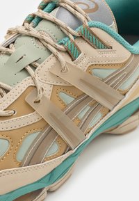 Scarpa atletica beige con accenti color turchese, dotata di una tomaia in rete testurizzata, lacci spessi e sovrapposizioni in plastica per supporto e design.
