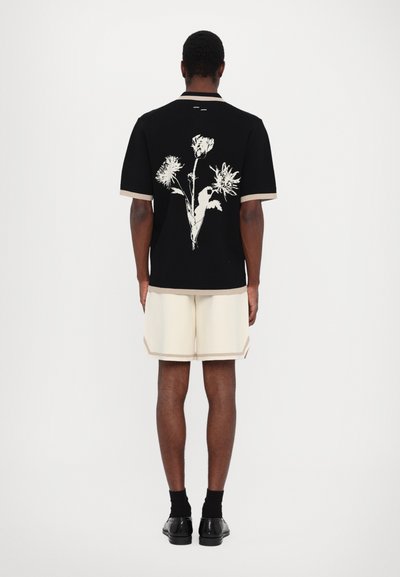 Schwarzes Kurzarmshirt mit einem weißen Blumenmuster auf dem Rücken. Kombiniert mit cremefarbenen Shorts und schwarzen Schuhen. Glatte Textur.