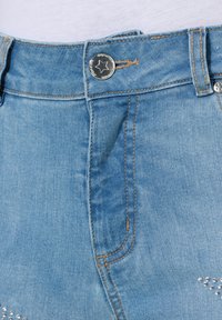 Pantalones cortos de mezclilla azul claro con un botón de presión plateado y costuras marrones. Tachuelas plateadas decorativas embellecen la parte inferior del frente.