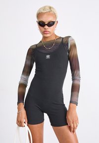 Černý žebrovaný overal s dlouhými průhlednými rukávy, které mají gradientový design. Produkt má na hrudi logo Adidas a je doplněn slunečními brýlemi.