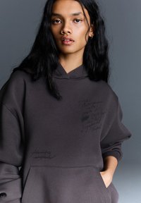 Hoodie gris foncé avec une poche kangourou, ornée d'accents d'écriture texturée dans diverses teintes. Le matériau semble doux et d'une coupe décontractée.