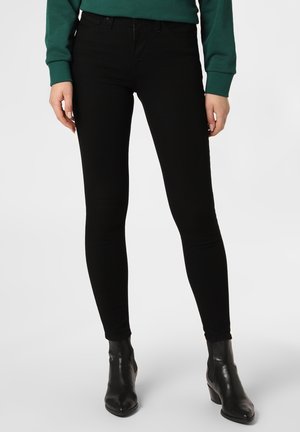 Personne portant un jean skinny noir, des bottines noires et un haut vert à manches longues, debout sur un fond blanc.