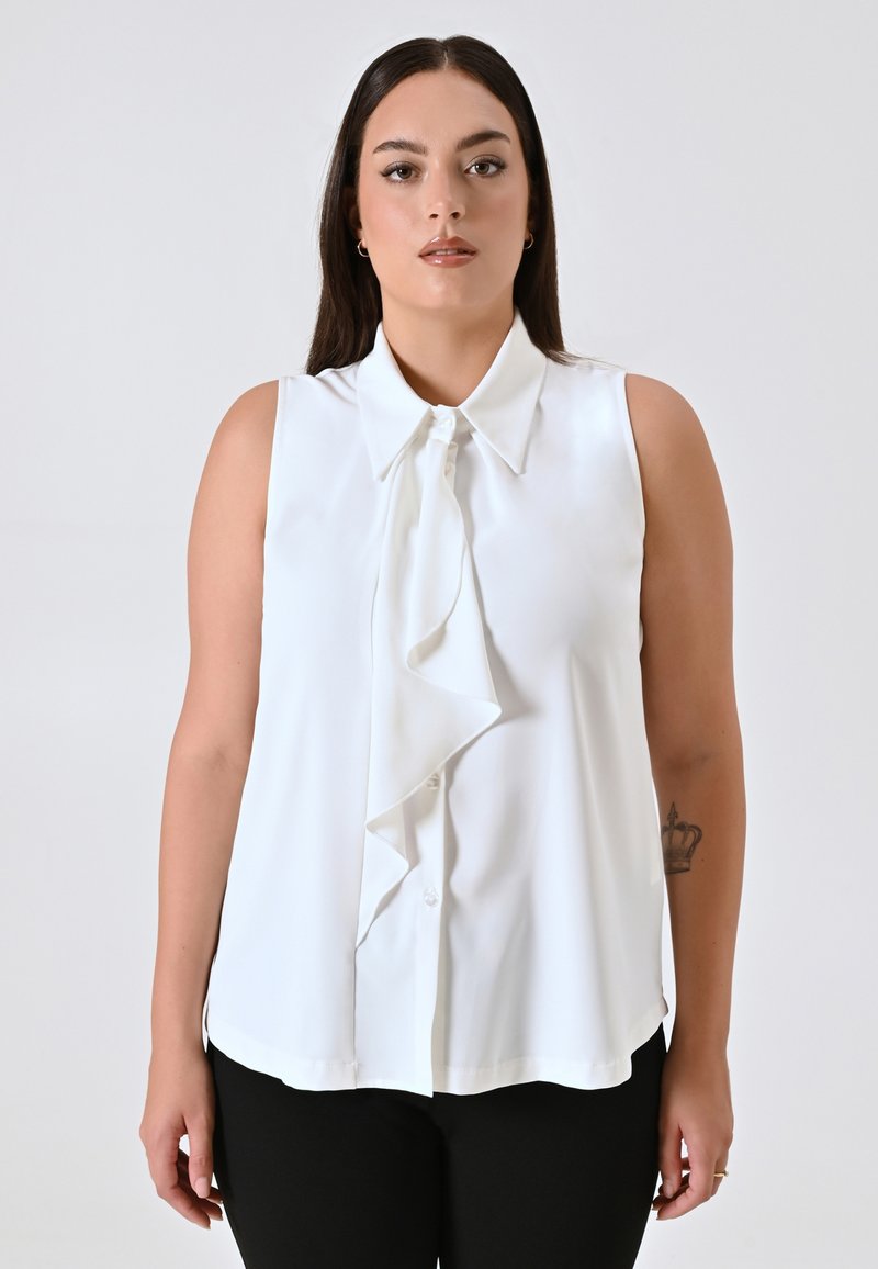 Blusa bianca senza maniche con un davanti arricciato, chiusura a bottoni e colletto. Tessuto morbido con una vestibilità comoda, abbinata a pantaloni neri.