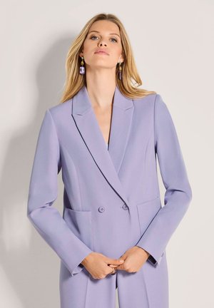 Lila double-breasted blazer met getailleerde revers, twee voorzakken en een gladde textuur. Model houdt blazer bij de taille vast, haar los.