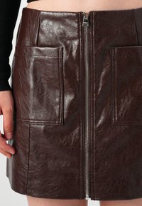 Mini gonna in pelle marrone con chiusura frontale a zip, due ampie tasche a toppa e una finitura sleek e testurizzata.