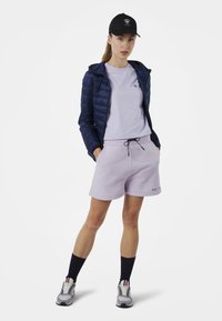 Giacca a vento blu marino sopra una t-shirt lilla, abbinata a pantaloni corti lilla. Cappellino nero, calzini blu marino e scarpe grigie con dettagli colorati.