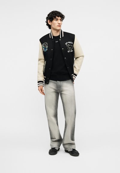 Νέος άνδρας που φοράει μαύρο και μπεζ varsity μπουφάν με κονκάρδες, μαύρο πουλόβερ, ανοιχτόγρι τζιν και μαύρα αθλητικά παπούτσια, στέκεται σε λευκό φόντο.