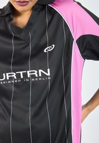 Zwarte sporttrui met roze accenten, verticale witte strepen en een kraag. Bevat logo en de tekst "ONTWORPEN IN BERLIJN" duidelijk weergegeven.