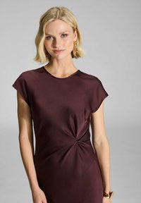 Burgundy mekko, jossa on hihat, sileä pinta, kierretyt yksityiskohdat vyötäröllä ja pyöreä pääntie. Yksinkertainen, napakka siluetti. Kultaiset koruaksentit.