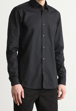 Mann trägt ein eng anliegendes, langärmeliges schwarzes Hemd mit Knöpfen und schwarze Hose, steht vor einem schlichten hellen Hintergrund.