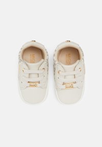 Zapatillas beige con un upper de tela texturizada y acentos de cuero liso. Presentan una placa con el logo dorado y cordones elásticos para un fácil uso.