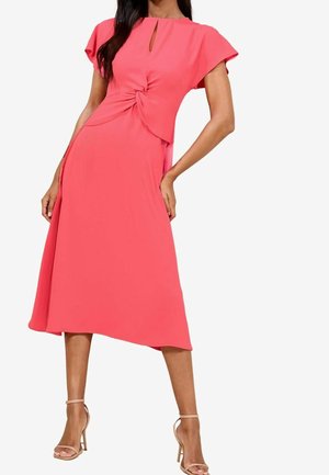 Femme portant une robe midi rose corail avec un détail de nœud torsadé à la taille et des manches courtes, associée à des talons nude à bride de cheville.
