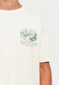 Hvit T-skjorte med grønne fugl illustreinger og rødt tekst som leser "The Gant Birding Club, New Haven Connecticut, Etablert 1949" på brystet.