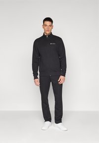 Schwarz Tracksuit aus Baumwollmischgewebe; Reißverschlussjacke mit hohem Kragen, Logodetail auf der Brust; eng anliegende Hose, kombiniert mit weißen Sneakern.