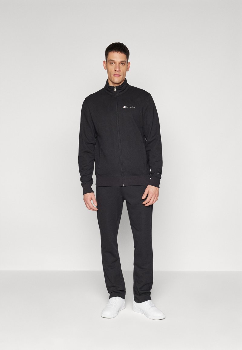 Schwarz Tracksuit aus Baumwollmischgewebe; Reißverschlussjacke mit hohem Kragen, Logodetail auf der Brust; eng anliegende Hose, kombiniert mit weißen Sneakern.