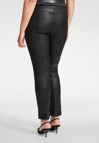 Zwarte broek van imitatieleer met een nauwsluitende pasvorm, voorzien van naden aan de achterkant en een gladde textuur. Draagt zwarte sandalen met hoge hakken en bandjes.