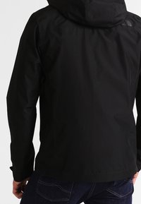 Schwarze wasserdichte Jacke mit Kapuze, die eine glatte Textur, ein minimalistisches Design und ein Logo auf dem oberen Rücken aufweist.