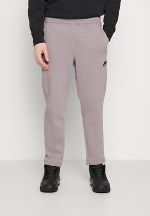 Personne portant un pantalon de survêtement Nike mauve clair avec logo noir, des baskets noires et un haut noir à manches longues, debout devant un fond uni.