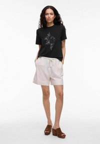 Kvinna med långt lockigt hår som bär en svart grafisk t-shirt, ljusbeiga shorts med dragsko och bruna klackade sandaler med öppen tå, stående med händerna i fickorna.