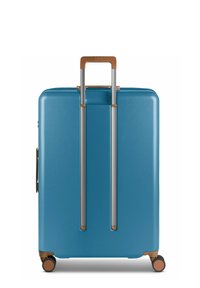 Maleta azul de carcasa dura con textura suave, que presenta un mango retráctil plateado y detalles en marrón; incluye cuatro ruedas giratorias.