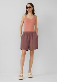 Lichtroze tanktop en losse, stofroze shorts met een elastische tailleband. Het model draagt een paar bruine zonnebrillen en beige platform sandalen.