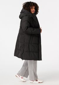 Zwarte pufferjas met capuchon, met een iets oversized pasvorm, gewatteerde textuur en zijzakken. Gecombineerd met grijze wijde broek en sneaker.