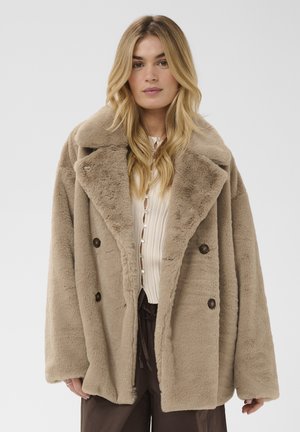 Blonde Frau trägt einen beige oversized Kunstpelzmantel, weißes knopfgeschlossenes Oberteil und dunkle Hose, steht vor einem schlichten hellen Hintergrund.