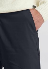 Hand met een crèmekleurige gebreide trui, waarbij de arm gedeeltelijk in de zak van de donkerblauwe broek zit, waarbij de stoftextuur en de stiksels zichtbaar zijn.