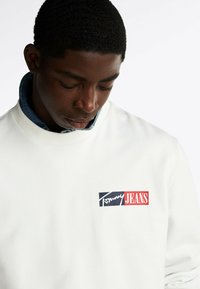 Weißer Pullover mit Rippkragen, versehen mit einem kleinen Logo, das "Tommy" in Blau und "JEANS" in Rot auf der linken Brust zeigt.