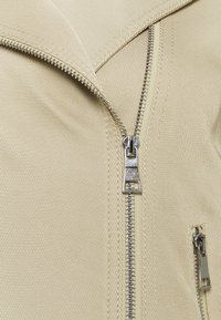 Chaqueta beige de cremallera con un amplio cuello, que presenta una cremallera plateada prominente y una textura de canalé fina. Bolsillo adicional con cremallera en el lateral.