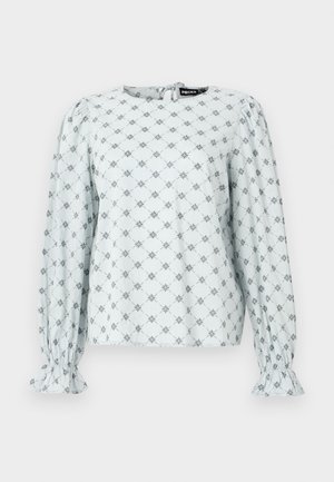 Blouse bleu clair avec de longues manches bouffantes, arborant un motif floral en losange de couleur marine. Manches élastiquées et fermeture au dos en forme de trou de clé.