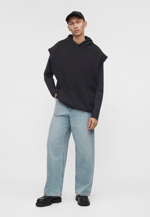 Jeune homme portant un sweat à capuche sans manches noir sur un t-shirt à manches longues, jean bleu clair, casquette noire et chaussures noires, debout les mains dans les poches.