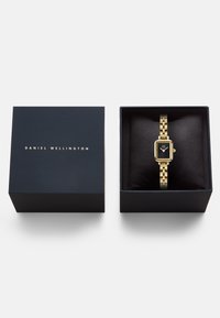 Gold-tone watch with rectangular face and black dial, displayed on a cushion inside a navy blue box with gold logo lettering.
