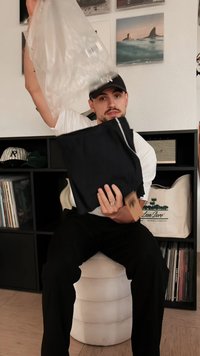 Musta vaatekappale pidetään yhdessä kädessä, muovipussi toisessa. Siinä on etiketti, sileä tekstuuri ja taiteltu muoto. Taustalla näkyy hyllyjä.