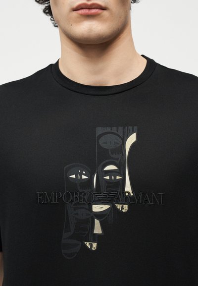 Κοντινό του σώματος που φοράει μαύρο μπλουζάκι με στυλιζαρισμένα αφηρημένα πρόσωπα και το λογότυπο "Emporio Armani" στο κέντρο του στήθους.