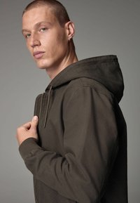 Hanorac de culoare verde măsliniu închis, cu șnur ajustabil, închidere cu fermoar și manșete ribbate. Textură moale, cu o croială lejeră și un design minimal.