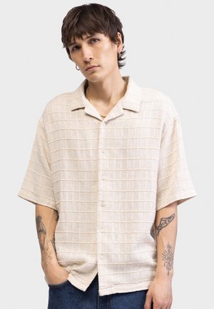 Ung mann med kort mørkt hår iført beige teksturert skjorte med knapper og gullkjede, viser tatoveringer på begge underarmer, hendene i lommene.