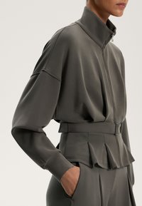 Siva zip-up jakna z visokim ovratnikom, dolgimi rokavi in poševnimi žepi; vsebuje pas in nabrano podrobnost na robu.